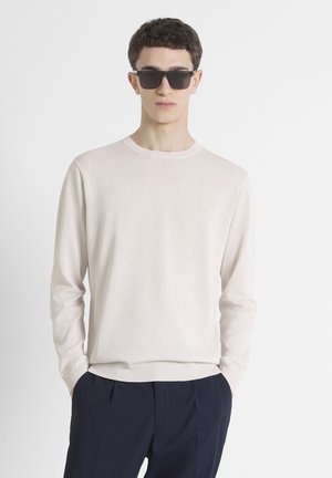 Beige gebreide trui met lange mouwen en ronde hals, met een subtiele logo op de borst, gedragen met marineblauwe broek en donkere zonnebril.
