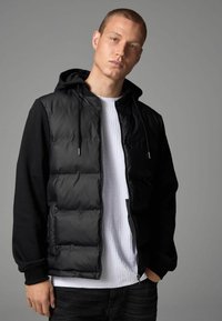 Doudoune noire avec fermeture éclair à l'avant, capuche intégrée et manches noires. Portée par-dessus une chemise texturée blanche. Le mannequin se tient contre un fond gris.