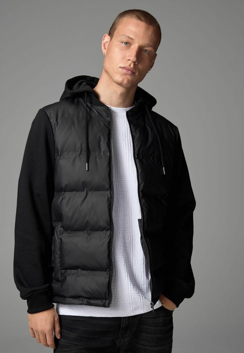 Doudoune noire avec fermeture éclair à l'avant, capuche intégrée et manches noires. Portée par-dessus une chemise texturée blanche. Le mannequin se tient contre un fond gris.