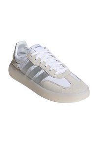 Baskets blanches avec des accents en suède gris, trois rayures grises, et une semelle lisse et arrondie. Les lacets sont blancs, et la chaussure dispose d'un col rembourré.