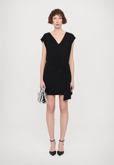 Robe noire courte avec un décolleté en V et des manches courtes, présentant un détail drapé sur le côté. Associée à un sac à main argenté et des chaussures à talons noirs.