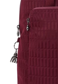 Bordeaux ryggsäck med texturerat tyg, som har en dragkedja ficka och en dekorativ svart charm. Betonas av glänsande silverfärgad metall.