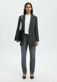 Mujer con cabello largo y oscuro que lleva una chaqueta gris oscuro, camiseta blanca, jeans grises, zapatos negros puntiagudos y lleva un bolso negro de hombro.
