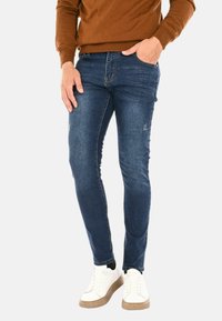 Jeans di denim di colore blu scuro con vestibilità slim. Presentano un leggero effetto usurato e un design a cinque tasche. Abbinati a sneakers bianche.