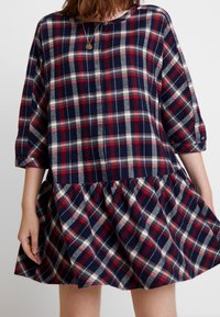 Femme portant une robe ample à carreaux rouge, bleu marine et blanc, avec des manches trois-quarts et un ourlet à volants, assortie à un collier pendentif rond.