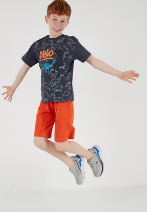 Ragazzo dai capelli rossi che salta, indossa una maglietta scura con stampa di dinosauri, pantaloncini arancioni vivaci e scarpe da ginnastica grigie con dettagli blu su uno sfondo semplice.