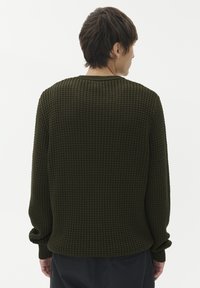 Vīrietis, kurš stāv ar muguru, ģērbies tumšzaļā teksturētā adītā džemperī un tumšās biksēs pret vienkāršu baltu fona.