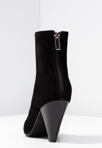 Czarne zamszowe botki za kostkę z wysmukłą, wysoką szpilką, zamkiem błyskawicznym z boku i gładką fakturą. Widok z tyłu ukazuje smukły kształt i projekt obcasa.