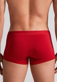 Shorts de boxe rouges en tissu doux, offrant une coupe ajustée et une taille élastique avec un logo. Texture lisse et design minimaliste.