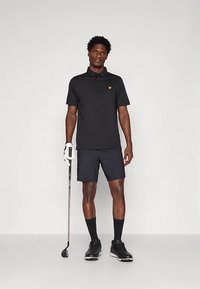 Άνδρας ντυμένος με μαύρη γκολφ εμφάνιση, κρατώντας ένα γκολφ μπαστούνι με γάντι στο αριστερό του χέρι, στέκεται μπροστά σε απλό φωτεινό φόντο.