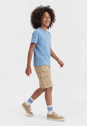 Niño sonriente caminando, con camiseta azul claro, pantalones cortos cargo beige, calcetines a rayas y zapatillas beige sobre fondo liso.