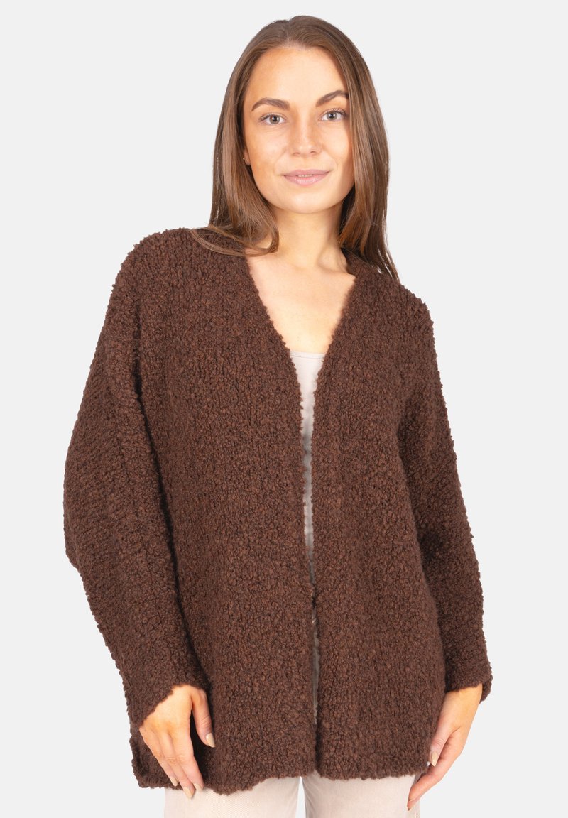 Femme portant un cardigan beige à la texture lâche sur un top et un pantalon clairs, devant un fond gris clair uni.
