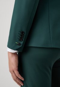 Veste de costume taillée vert foncé avec une texture lisse, quatre boutons noirs sur les poignets et un poignet de chemise blanche visible en dessous.