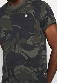 Camiseta de manga corta con estampado de camuflaje en verde oscuro y gris. Hecha de algodón suave, tiene un cuello redondo y mangas enrolladas. Logo en el dobladillo del pecho.