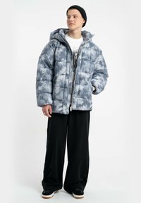 Padded szara i biała kurtka tie-dye z kapturem, z zamkiem błyskawicznym z przodu i prostym krojem. Połączona z czarnymi spodniami o szerokich nogawkach i sneakersami.