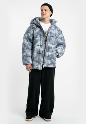 Padded szara i biała kurtka tie-dye z kapturem, z zamkiem błyskawicznym z przodu i prostym krojem. Połączona z czarnymi spodniami o szerokich nogawkach i sneakersami.