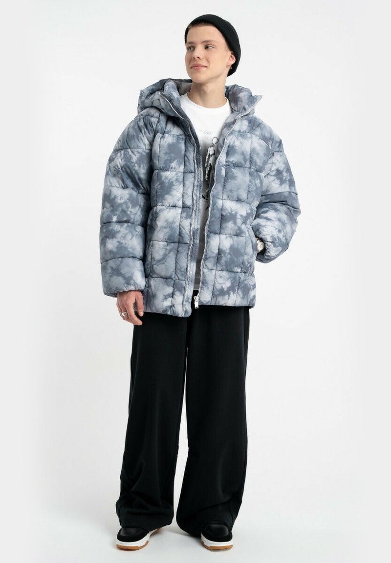 Padded szara i biała kurtka tie-dye z kapturem, z zamkiem błyskawicznym z przodu i prostym krojem. Połączona z czarnymi spodniami o szerokich nogawkach i sneakersami.