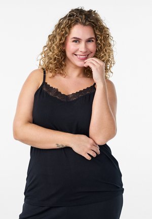 Naeratav naine, lokkis juustega, kandes musta ilma varrukateta pitsäärisega ülaosa, poseerides käega näo lähedal valge tausta ees.