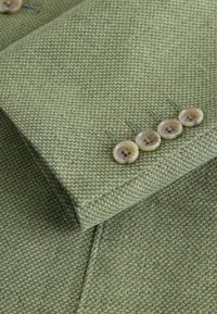 Gros plan sur la manche d’un blazer texturé vert olive avec quatre boutons beiges cousus en rangée près du poignet.
