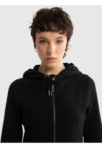 Tânăr cu păr scurt, creț și întunecat, purtând cercei mici inelari și o hanorac negru cu fermoar, pe un fundal simplu, deschis la culoare.