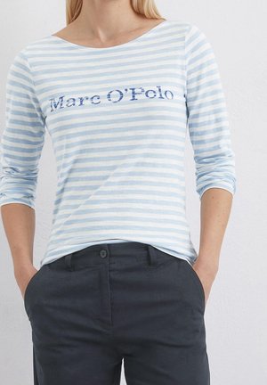 Femme portant une chemise à manches longues rayée bleu clair et blanc avec le texte "Marc O'Polo" et un pantalon bleu foncé, les mains dans les poches.