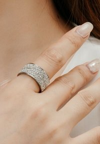Anello d'argento con due fasce texturizzate, una liscia e una decorata con gemme trasparenti, indossato su un dito con unghie smaltate.