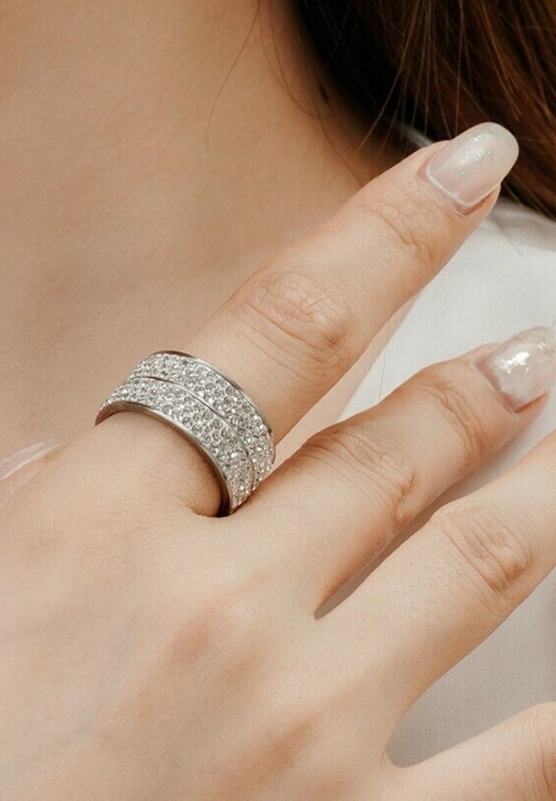 Anello d'argento con due fasce texturizzate, una liscia e una decorata con gemme trasparenti, indossato su un dito con unghie smaltate.
