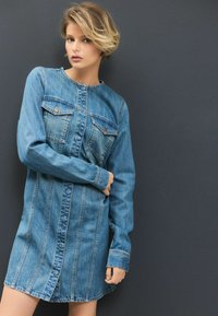 Denim overhemdjurk in lichtblauw met lange mouwen, knoopsluiting aan de voorkant, twee borstzakken en verticale stiksels.