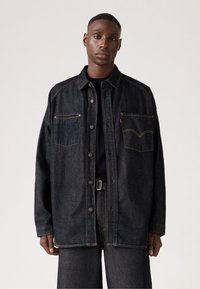 Donkerdenimjack met knoopsluiting, twee borstzakken en contrasterende gele stiksels. Vrije pasvorm, lange mouwen en een rechte onderzoom.
