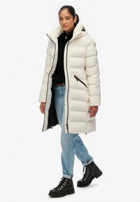 Witte, gewatteerde lange jas met kap, voorzien van zwarte ritsen en biezen, gedragen over een zwarte trui en lichtblauwe spijkerbroek met zwarte laarzen.