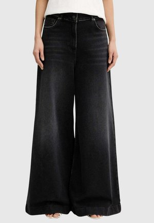 Donna con jeans neri a gamba larga e top bianco, piedi visibili con sandali aperti, in piedi davanti a uno sfondo uniforme.