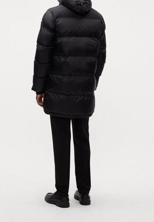 Osoba w długiej czarnej kurtce puchowej z kapturem, czarnych spodniach i czarnych butach, stojąca tyłem na jednolitym tle.