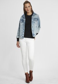 Jachetă din denim cu o spălare deschisă, pulover texturat negru, blugi albi skinny și botine maronii până la gleznă. Design de ținută casual în straturi.