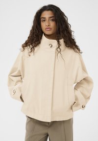 Béžna bombažna jakna z visokim ovratnikom in zapiranjem na gumbe. Ima velike stranske žepe in široke rokave z detajli z gumbi.