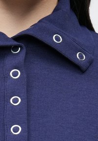 Top bleumarin cu dungi, prevăzut cu un guler pliat cu inele argintii și închidere cu nasturi. Materialul pare texturat și elastic.