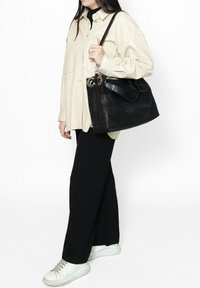 Borsa a tracolla nera in pelle con superficie texturizzata, grande hardware circolare argentato e manico nero a contrasto. Indossata con una camicia oversize di colore chiaro.