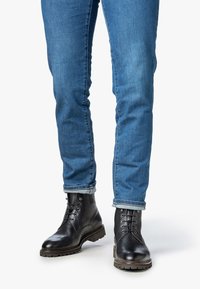 Persona con jeans azules arremangados y botas de cuero negras con cordones, de pie sobre una superficie blanca.