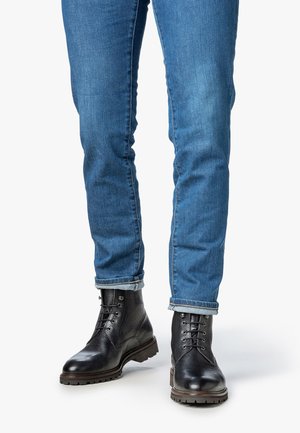 Persona con jeans azules arremangados y botas de cuero negras con cordones, de pie sobre una superficie blanca.
