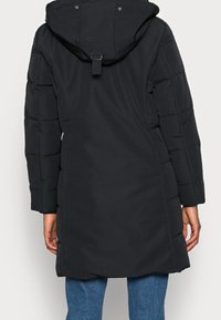 Sort polstret jakke med hætte, lange ærmer og indsnævret talje. Har en skjult lynlås og trykknapper. Glat, mat tekstur.