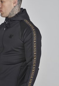 Schwarze Zip-Hoodie aus strukturierter Stoff, mit einem goldenen Logo am Ärmel und einer verstellbaren Kapuze. Eng anliegendes Design.