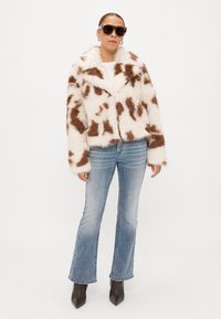 Casaco de pelo falso com uma base branca e manchas castanhas, apresentando um colarinho e mangas longas. Combinado com jeans flare azuis e botas de tornozelo.