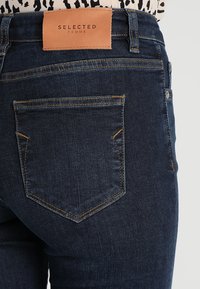 Un bolsillo trasero de unos vaqueros de mezclilla azul oscuro con costuras naranjas y una etiqueta de marca de cuero marrón claro que dice "SELECTED FEMME" en la cinturilla.
