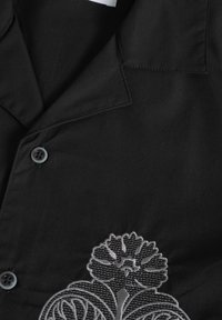 Camisa negra de botón con collar, textura de tela y elaborados bordados florales plateados en el lado inferior izquierdo.