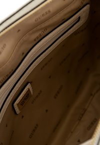 Interior beige de un bolso, con una textura de tela suave que presenta sutiles logotipos de "GUESS" y una etiqueta metálica de la marca. Comparto con cremallera visible.