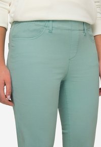Pantalones verde claro hechos de una tela suave y elástica, con bolsillos frontales, trabillas para el cinturón y costuras en contraste para un detalle adicional.