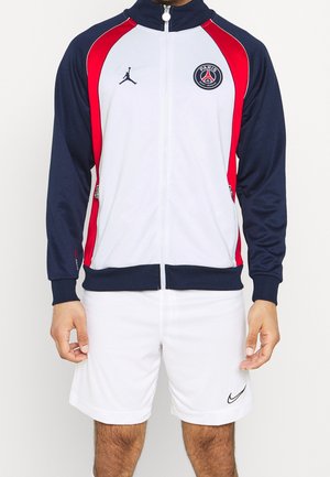 Personne portant une veste de sport Paris Saint-Germain blanche, marine et rouge et un short blanc Nike, debout devant un fond uni.