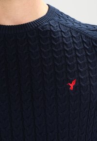 Pull en maille bleu marine avec un design en câble texturé, doté d'un col rond et d'un petit logo d'oiseau rouge sur la poitrine.