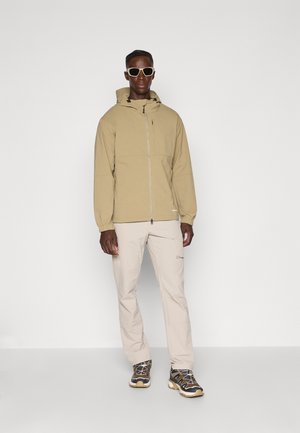 Veste à capuche beige avec fermeture éclair à l'avant, dotée d'une poche poitrine. Associée à un pantalon beige clair et des baskets noires avec des accents jaunes.