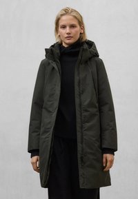 Tmavě zelená parka s kapucí, vybavená zipem a zapínáním na druky, vyrobená z hladké, odolné látky, nošená přes černý svetr.