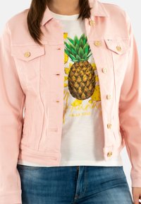 Chaqueta de denim rosa claro con botones dorados, que presenta dos bolsillos en el pecho y un diseño ajustado, llevada sobre una camiseta blanca con un gráfico de piña.